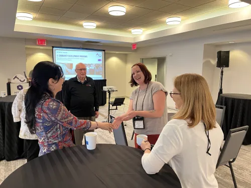 A smiling woman reaches her hand out to greet others in a meeting room