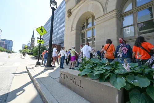 A group of people hold hands with heads bowed in prayer while facing a building. A sign in the foreground identifies it as City Hall.