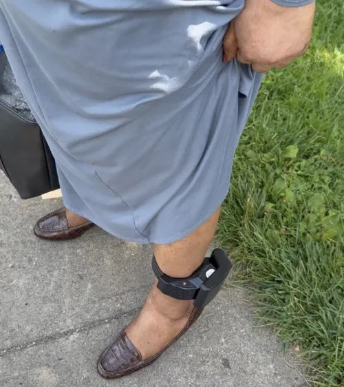 A woman's leg is visible, bearing an ankle monitor situated above a dress shoe. The woman holds up her dress to show the monitor.
