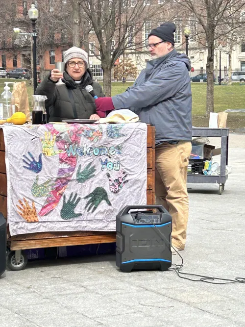 A woman in a puffy black coat holds up a silver chalice from behind a communion table. To her left, a man holds up a microphone for her to speak into. They are outside on the Boston Common.