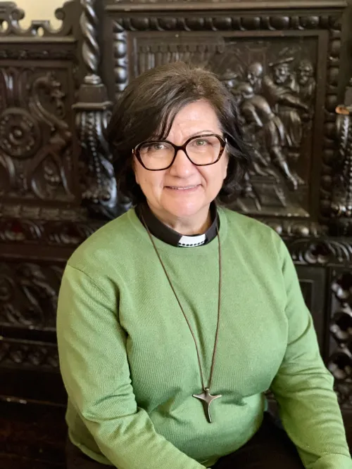A woman in a green sweater smiles at the camera for a headshot. Behind her is ornately carved dark wood panelling.