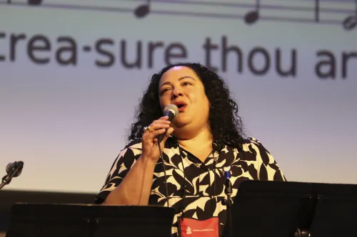 A woman with dark, curly hair in a white patterned blouse sings into a microphone with lyrics on a screen behind her.