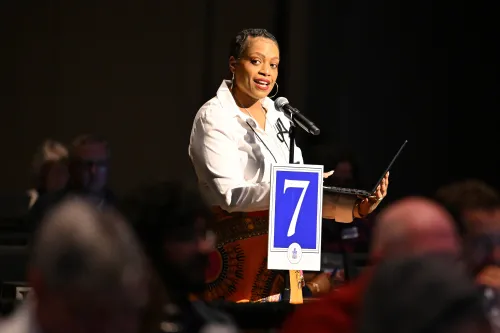 A woman in a white shirt speaks into a microphone and addresses a dark room  