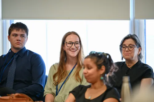 A young man, a young woman, and an older woman sit along a windowed wall, listening to a conversation. The young woman is smiling.