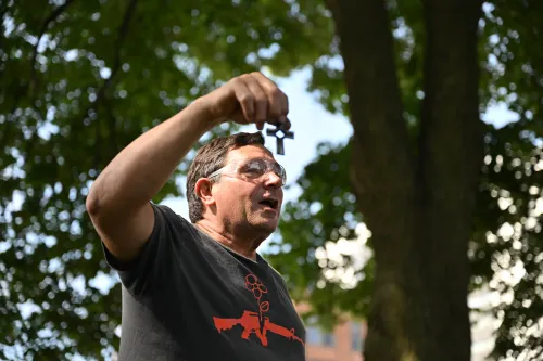 A white man in a dark gray shirt holds up a metal cross made from former firearms.