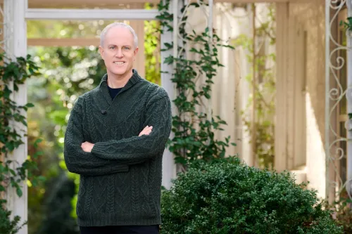 Man in sweater stands with hands crossed in an outdoor area with greenery