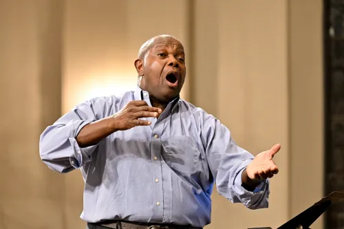 A man in a blue oxford shirt holds his arms out conducting an unseen choir
