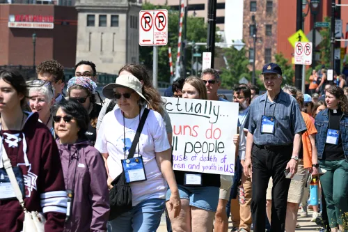 A variety of people wearing GA227 neck badges walk in unison. One holds a sign identified gun violence as the number cause of death for children in the US. 