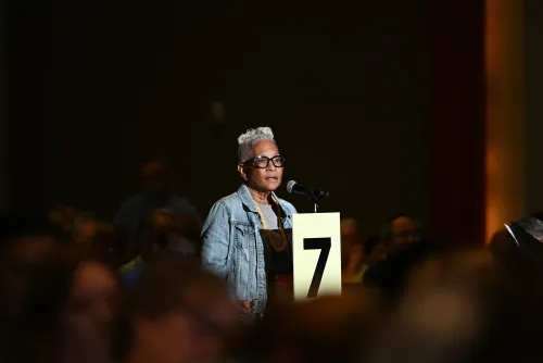A woman with short white hair in a denim overshirt stands at a microphone in a dark room, surrounded by a seated crowd