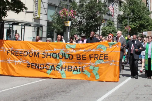 A group of people in clergy garb march, with a large orange banner that reads "Freedom should be free, end cash bail"