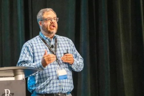 A man in a blue shirt speaks to an unseen audience.