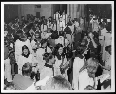 The interior sanctuary of The Church of the Advocate in Philadelphia is full from wall-to-wall with people. This photo was taken on July 29, 1974, at the controversial ordination ceremony of the Philadelphia Eleven. The eleven women ordained that day are seen in the image, wearing white robes and chatting amongst themselves as they move through the crowd.