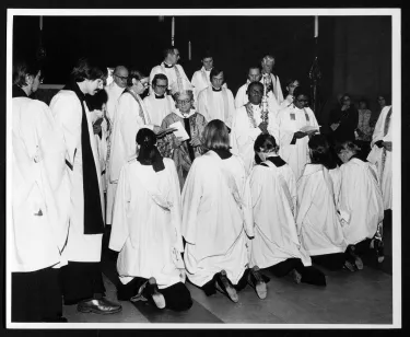 Five women in white robes kneel before a man wearing the robes of an Episcopalian bishop. He is Episcopal Bishop Paul Moore, Jr., and here he refuses to ordain as priests five women deacons presented to him during the ordination of five male deacons at the Cathedral of St. John the Divine in New York. He permitted the women, however, to go through every part of the service except the laying on of hands.