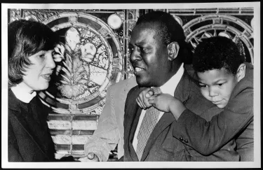 A photograph of two adults, one woman and one man, standing in front of a stained glass window. There is a boy clinging to the man's back. The woman wears a white robe; the Black man wears a suit. His son's hands are clasped below his chin, over his tie. He smiles at the woman.