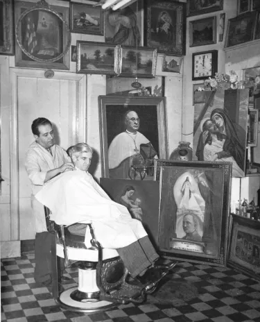 A man sits in a barber's chair as he gets his hair cut. The barber stands behind him. The walls and floor around them are covered with paintings, created by the barber's very hand.