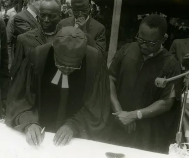 Black and white photograph of a man leaning over a table to sign a document, a pen gripped in his right hand. Signing of Integration papers at Luluabourg by Legal Representative Pastor Bakatushipa Pierre and Pastor Nsenda Joseph (Executive Secretary of the Church), March 1970.