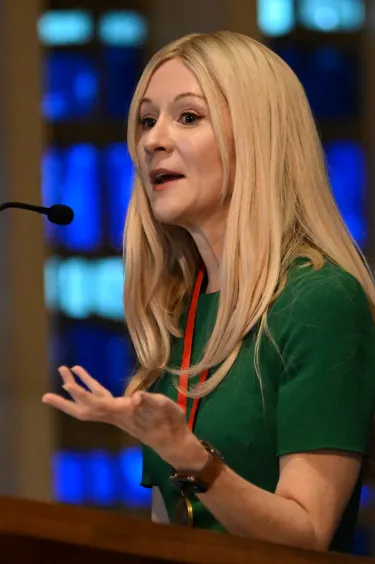 A close up of a woman at a podium speaking with her hand raised
