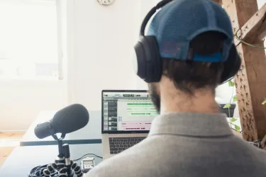 The back of a male podcaster's head is shown as he sits near a microphone and computer screen