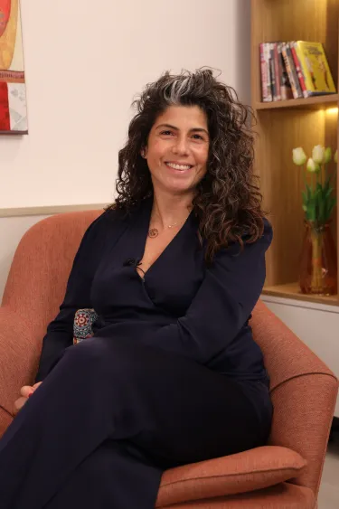 A smiling woman with wavy hair sits in a cinnamon colored chair near a book shelf.