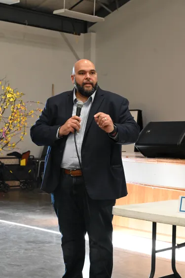 A man in a dark blazer speaks while holding a microphone.