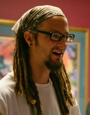 A man with long braids tied back with a bandana speaks to someone unseen in a dark room. 