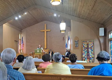 The sanctuary of a church. People sit in pews, with their backs to the camera.