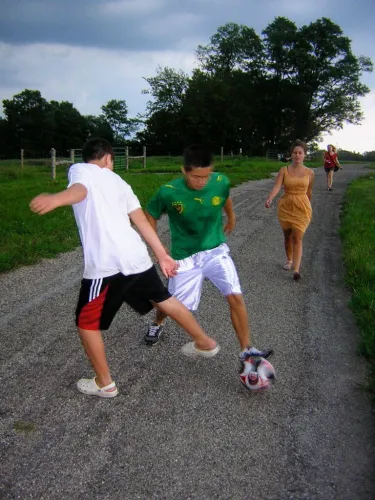 Two boys dribbling soccer ball with two others walking behind them down a empty road at Fox Hollow Farm