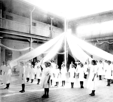 A group of perhaps twenty girls stand in a courtyard, with a May Day pole at the center. The girls all wear white.