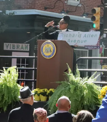 Rev. Cavaness of Mother Bethel stands on stage holding up an old sign that reads "Richard Allen Way." She is telling the crowd the story of the sign.