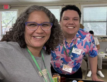 A curly haired woman in a gray shirt smiles next to a curly haired gentleman in a colorful pink and blue shirt.