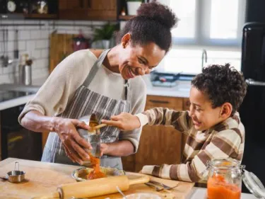 A little boy helps his mother in the kitchen by scooping food from a mixing bowl into a dish.