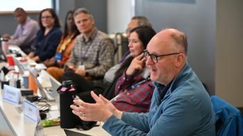 A man speaks at a table with other people in the background sitting at a table with him.