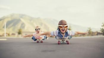 A young boy and girl are wearing flying goggles while outstretching their arms to attempt flying while on skateboards. They have large smiles and are imagining taking off into the sky.