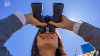 a woman with long dark hair looks into a large pair of black binoculars while smiling