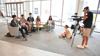 A videographer and interviewer talk with a group of five people seated on a couch in the lobby of a large building.