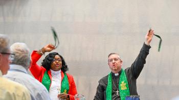 A man and woman in clerical garb wave palm leaves in the air at a communion table.