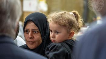 A woman with her head covered carries a young girl who has golden hair tied up into a ponytail
