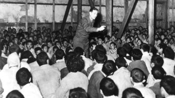 Harold Voelkel is seen standing in the middle of a crowded room, with his right arm extended and his finger pointed in instruction. He is speaking to Korean POWs. 