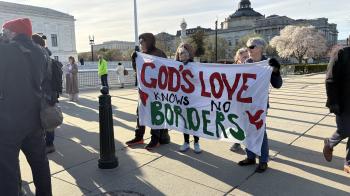 A group of faith leaders stand together holding a large white cloth banner between them. It says "God's love knows no borders" in red and green block text.