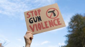 a hand holds up a brown cardboard sign against a blue sky. The sign says "stop gun violence" in red and black with an image of a gun surrounded by a red circle with a line through it.