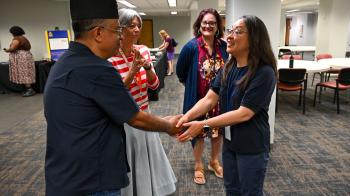 A smiling woman shakes hands with a man wearing a hat while others look on.