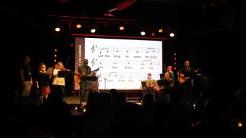 Musicians play a song in a bar with the words behind them on a projector screen