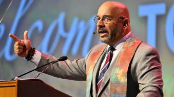 A man in a light-colored suit and colorful stole speaks at a lectern. 
