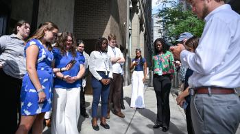A group of young people pray, some looking at the prayer on their smartphones, in front of a building in New York City.