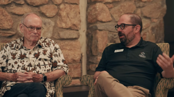 Two men casually attired talk in front of a stone wall