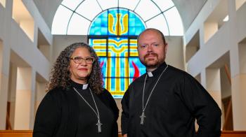 A man and a woman stand side by side in clergy collars and black shirts with silver cross necklaces. A stained glass window is behind them.