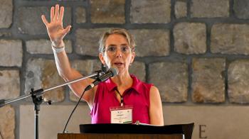 A person in a pink top stands at a podium with a microphone, raising one hand while speaking, against a stone wall backdrop.