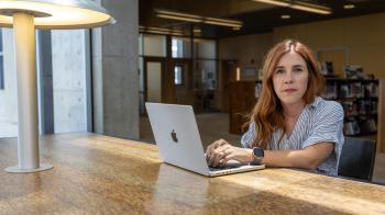 A person with long reddish hair sits at a wooden table in a library, typing on a laptop under the glow of a modern lamp.