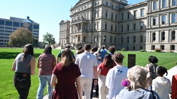 a group of several dozen young adults walk towards the state capital building in Lansing, MI