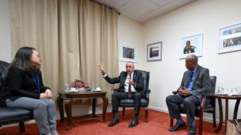 A Cuban man in a business suit sits in a chair and  talks while gesturing with one hand raised. On either side of him, an Asian woman and a Black man, both in business attire, lean in to listen.